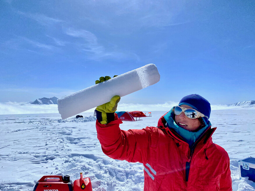 A woman dressed in winter clothing is surrounded by snow and ice on a sunny day holds up an ice core.