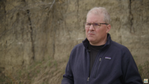Man being interviewed in front of a sandy riverbank.