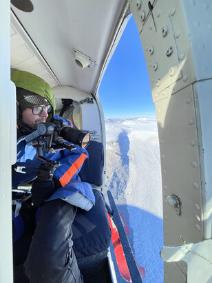 Man sitting in a helicopter with the door open with snow capped mountains in the outdoor background.