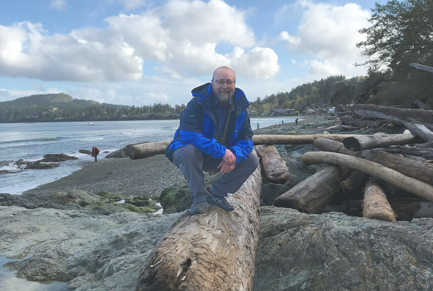 Man squatting on a log with arms crossed against a background of the ocean.