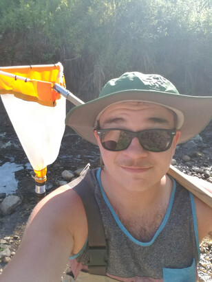 Young man with sunglasses on takes a selfie holding a net with a stream in the background.