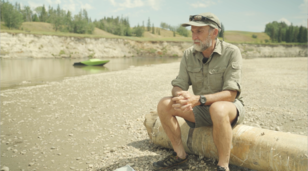 Man sitting on a log beside a river being interviewed on a sunny day.