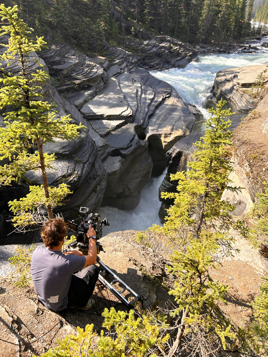 Man sits above waterfalls filming with a movie camera.