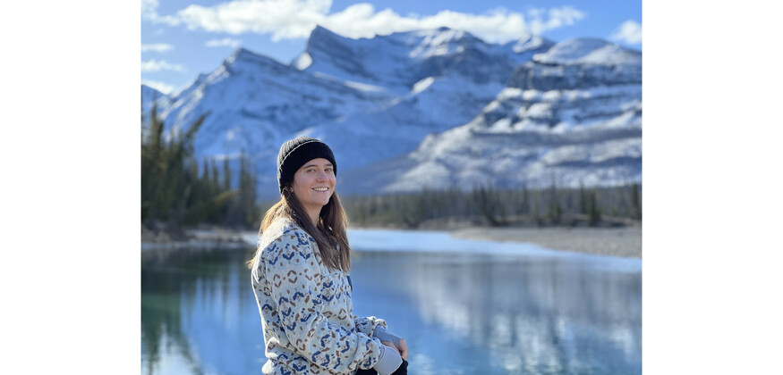 Young woman with long hair and wearing a sweater stands in front of mountains and a lake. Photo credit: Ryan Northcott.