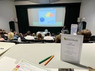View over crowd with desk in foreground and projector screen in the background with a map of the North Saskatchewan River Watershed.