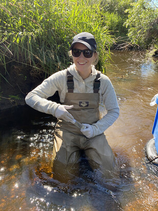 Young woman kneeling in waders and a tube in a shallow stream.