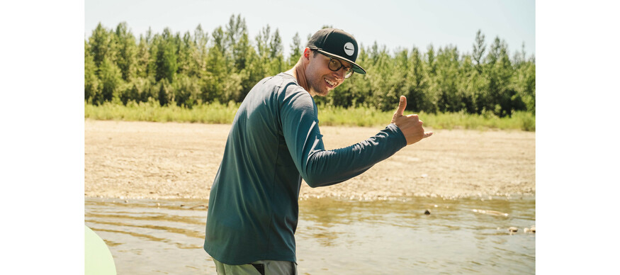 Man gives shaka sign to the camera while wading out of a river on a sunny day.