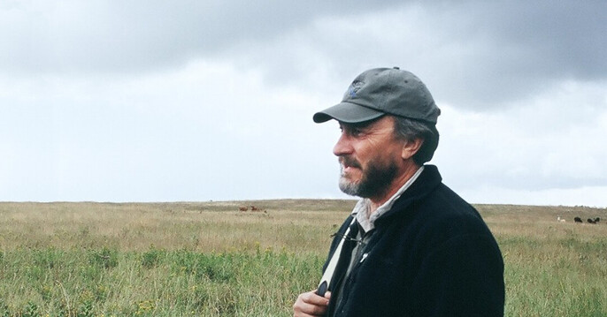 Fitch standing in a field talking to someone outside the shot with cows grazing in the background.